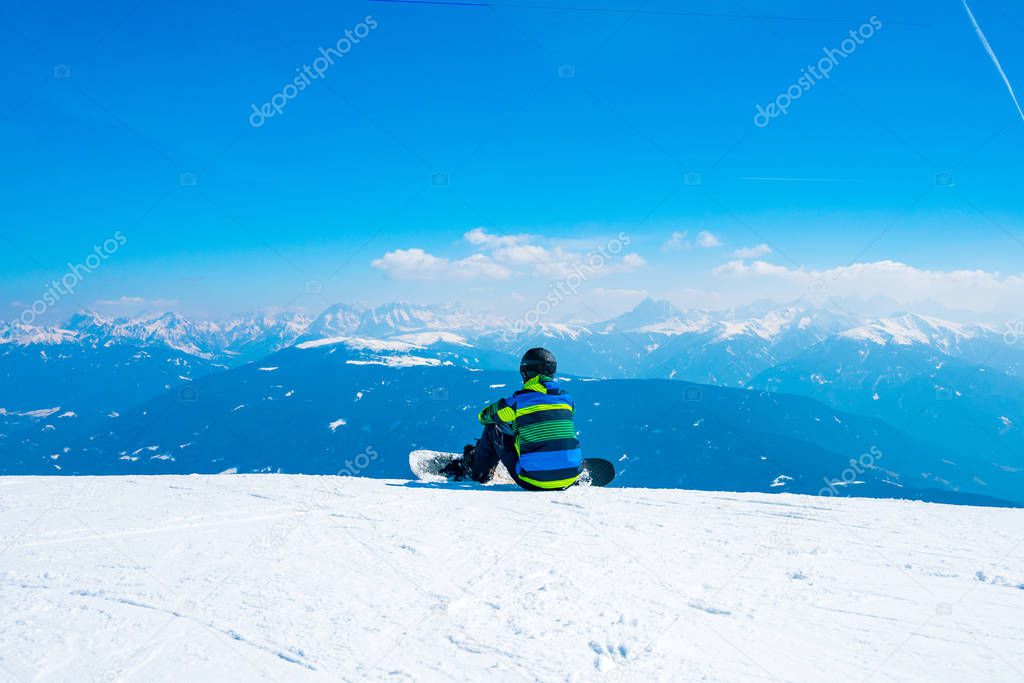 Saalbach, Austria. March 20, 2018. young with a snowboard man sitting on the top of the mountain in Alps watching into the horizon. Admiring ski resort. Snowboarding.