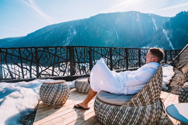 Genç adam kış süre içinde inanılmaz bir dağ manzaralı spa açık bir teras üzerinde tüyler ürpertici bir elbise içinde. Kayak Merkezi.