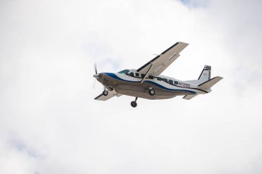 Mavi ve siyah çizgili beyaz bir uçak bulutlu bir gökyüzünün altında havada uçuyor. Pervane ve genişletilmiş iniş takımları görünüyor..