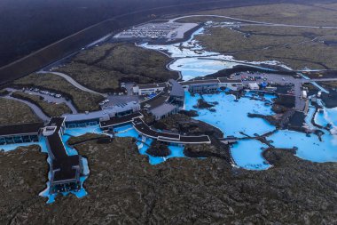 Blue Lagoon Spa 'nın süt mavisi havuzları, yakınlardaki otel binaları, yollar ve olay yerinde görülebilen bir otopark ile koyu lav tarlalarıyla tezat oluşturuyor..