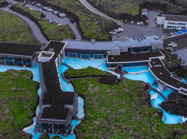 Mavi Göl Kaplıcaları 'nın turkuaz jeotermal havuzu karanlık lav tarlaları boyunca esiyor, modern otel binaları ve kısmen tıkanmış yollar görülebilir..