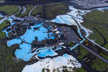 Blue Lagoon Spa 'nın sütlü mavi havuzları koyu lav tarlalarıyla, otel, park alanı ve kısmen tıkanmış yollarla tezat oluşturuyor..
