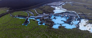 Mavi Göl Kaplıcaları 'nın turkuaz jeotermal havuzları karanlık lav tarlalarıyla çevrili, modern binalar ve kısmen işgal edilmiş yollar..