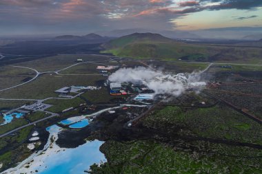 Blue Lagoon Spa 'nın mavi jeotermal suları koyu lav tarlaları, yükselen buhar, yakındaki otel ve uzak yeşil tepeler ve dağlarla tezat oluşturuyor..