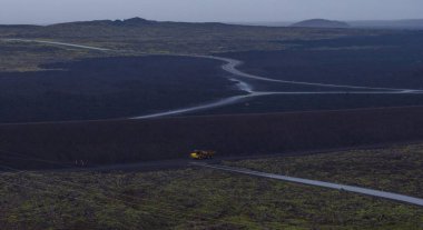 Yeşil yosun yamaları ve bulutlu gökyüzünün altında sarı bir inşaat aracıyla İzlanda 'da karanlık bir lav tarlasından geçen dolambaçlı bir yol..