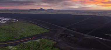 İzlanda 'daki kıvrımlı bir yol, elektrik hatları, yeşil yosun bantları, kırmızı bir araç ve bulutlu bir gökyüzünün altındaki uzak tepelerin havadan görünüşü..