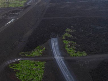 İzlanda 'da koyu lav alanları ve yeşil yosunlarla çevrili bir yol kesişiminin hava görüntüsü, elektrik hatları ve kullanım amaçlı yapılar görünür..