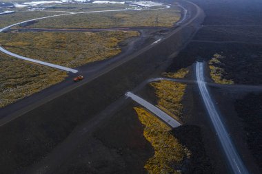 Koyu lav alanları dolambaçlı yollarla kesişiyor, biri turuncu bir araçla, İzlanda 'daki Mavi Göl' ün yanında, jeolojik aktivite gösteriyor..