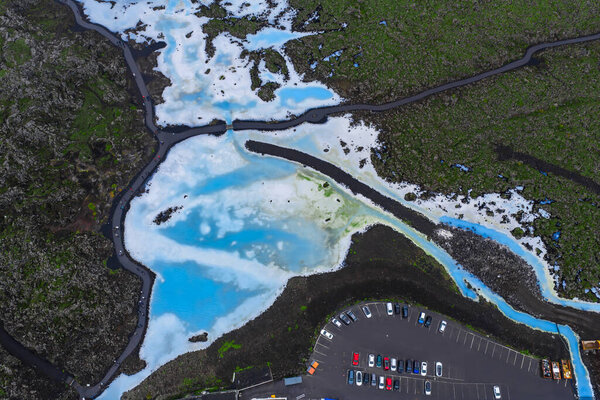 Голубые геотермальные воды Blue Lagoon Spa контрастируют с темными полями лавы, с дорогами, парковкой и видимой ниже активностью посетителей..