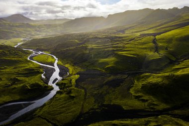 Sarmal bir nehir İzlanda 'da yeşil bir vadi boyunca akar, tepeler ve volkanik topraklarla çevrilidir, güneş ışığı ve gölgeler bulutlu bir gökyüzünün altında oynar..
