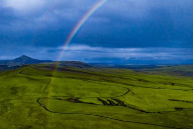 Bereketli yeşil arazi, dolambaçlı kara yollar, gökyüzünde canlı bir gökkuşağı, İzlanda 'da ıssız bir dağ zirvesi, ve bulutlu bir ufkun altında uzak bir su..