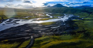 Sarmal bir nehir siyah volkanik kum ve yeşil yosunların arasından akar, arka planda tepeler ve dağlar İzlanda 'da parlayan günbatımı gökyüzünün altında.