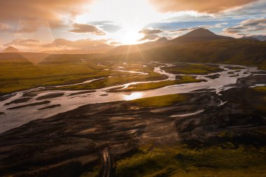 Siyah volkanik kum ve yeşil yosunlar arasında süzülen nehirlerin hava görüntüsü, İzlanda 'da gün batımında parlayan bir gökyüzüne karşı dağ silueti ile çevrili..