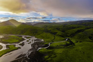 Yeşil yosun kaplı tepeler, siyah kum nehir yatakları ve altın günbatımı ışığıyla aydınlatılmış bir dağ ile İzlanda manzarası..
