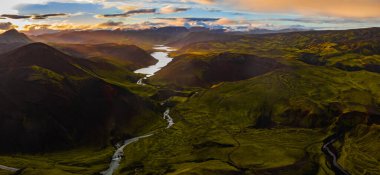 Yeşil bir vadide, volkanik tepeler ve kraterlerle çevrili dolambaçlı bir nehrin havadan görünüşü İzlanda 'da dağılmış bulutlarla altın bir günbatımının altında bir göle götürüyor..