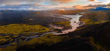 Gün batımında İzlanda 'nın hava manzarasında dolambaçlı bir nehir, yeşil yosun, volkanik toprak, uzak buzullar, kraterler ve bulutlu renkli bir gökyüzü var..
