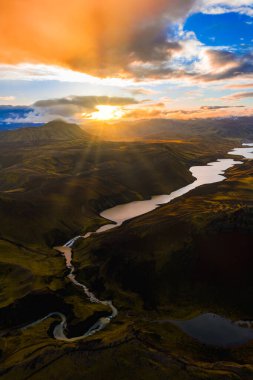 İzlanda 'nın yosunlu arazisinin göl, nehir ve tepeli havadan görünüşü. Altın günbatımı ışığı bulutların arasından süzülüp engebeli manzarayı aydınlatıyor..