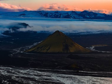 Karanlık ovalarda Maelifell volkanı şeklindeki bir koninin hava görüntüsü, beyaz nehir desenleri, buzul kaplı dağlar ve İzlanda 'da canlı bir günbatımı gökyüzü..