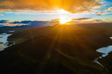 İzlanda 'nın engebeli arazisinin hava manzarasında volkanik kraterler, dolambaçlı bir nehir, dingin bir göl ve gün batımında dağınık bulutlu altın bir ışık var..