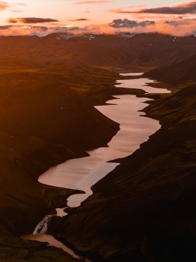 Koyu yeşil tepeler, küçük bir şelale, karlı zirveler ve İzlanda 'da ılık günbatımı renkleriyle çevrili bir yılan gölü..