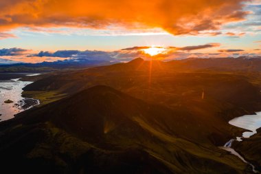 İzlanda 'nın volkanik tepelerinin, kraterlerin ve gün batımında dönen bir nehrin havadan görüntüsü. Karla kaplı dağlar ve dingin bir göl dramatik manzarayı güzelleştirir..