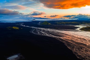 Güneş batarken İzlanda 'nın volkanik manzarasının havadan görünüşü, siyah arazi, buzul nehirleri, yeşil bir tepe ve canlı turuncu ve mavi bir gökyüzü içerir..