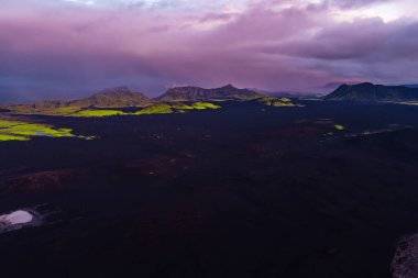 Yeşil yosun, engebeli dağ sırtları ve İzlanda 'da pembe ve mor bir gökyüzünün altında küçük beyaz bir göl ile karanlık volkanik ovaların dramatik hava manzarası..