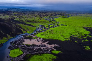 Karanlık volkanik topraklarla kaplı yeşil yosunların, dalgalı nehirlerin ve İzlanda 'da bulutlu bir gökyüzünün altındaki uzak tepelerin havadan görünüşü..
