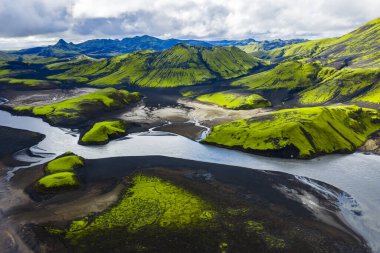 Yeşil yosun kaplı tepeler, karanlık volkanik toprak ve İzlanda 'da sarmal bir nehir, parçalı bulutlu bir gökyüzünün altında engebeli dağlar..