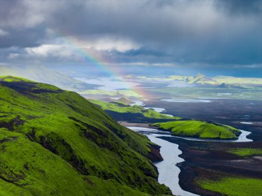 Yeşil tepeler, karanlık volkanik toprak, dolambaçlı bir nehir, soluk bir gökkuşağı ve ufuktaki engebeli araziyle İzlanda 'nın hava manzarası..