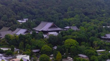 Japonya 'nın Kyoto kentindeki yoğun yeşil ormanlarla çevrili tarihi bir tapınağın serin hava manzarası. Kamera düzgün hareket ediyor, geleneksel mimariyi gösteriyor..