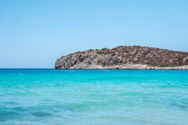 Kristal berrak turkuaz sular Girit 'in kıyı şeridine uzanan kayalık, çalılarla kaplı bir tepenin üzerinde canlı mavi bir gökyüzü ile karşılaşıyor..