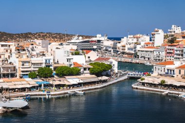 Yunanistan 'ın Girit kentindeki Agios Nikolaos kıyı kasabasından geçen su yolu gibi bir kanal, beyazlatılmış binalar, kırmızı kiremitli çatılar ve arka planda kenetlenmiş bir yolcu gemisi bulunuyor..