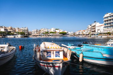 Yunanistan 'ın Girit kentindeki bir limanda küçük tekneler berrak mavi sularda yüzüyor. Beyaz ve bej renkli binaları olan bir liman kenti mavi gökyüzünün altında yer alır..