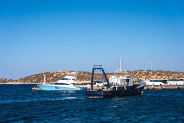 Yunanistan 'ın Girit kentindeki limanda beyaz bir yat ve mavi vinçli bir servis botu bulunuyor. Engebeli tepeler, park halindeki araçlar ve derin mavi sular görülebilir..