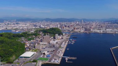 Japonya 'nın Hiroşima şehrinin havadan görünüşü, açık mavi gökyüzü altında bir yolcu gemisi, şehir yapıları, bir köprü ve dağların bulunduğu bir limanı gösteriyor..