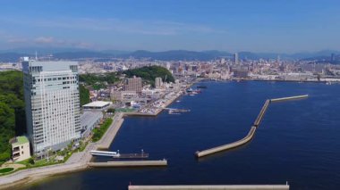 Japonya, Hiroşima 'nın havadan görünüşü, bir liman, modern yüksek binalar, yemyeşil alanlar ve arka planda dağlar olan kentsel binalar gösteriyor. Yavaş yavaş çevirme hareketi.