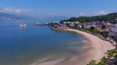 Mavi sularda ilerleyen feribotlarla Itsukushima Adası 'ndaki sahil manzarası, bir şehir manzarası, uzak dağlar ve kıyıdaki geleneksel binalar..