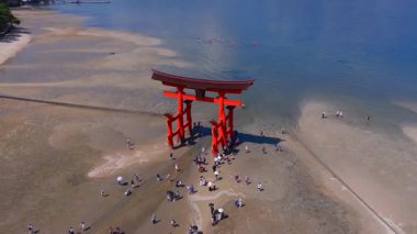 Itsukushima Adası 'ndaki ikonik kırmızı Torii kapısı. Dalgalar alçaldığında, ziyaretçiler kumsalda yürüyor ve mavi sularda kayakçılar hareket ediyor..