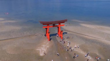 Itsukushima Adası 'ndaki ikonik kırmızı Torii kapısı sular çekildiğinde, ziyaretçiler kumsalda ve sığ sularda yürürken mavi gökyüzünü yansıtıyor..