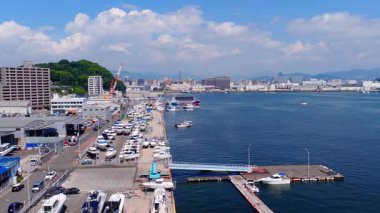 Hiroşima 'nın deniz manzarası sakin bir limanı, tekneleri, dalgaları, şehir yapılarını, yeşil tepeleri ve mavi gökyüzünün altındaki uzak dağları gösteriyor..