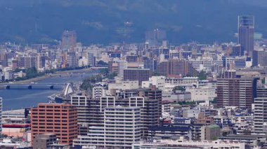 Japonya 'nın Hiroşima şehrinin hava manzarası, modern ve geleneksel binaları, şehir merkezinden akan bir nehri ve arka planda yeşil dağları gösteriyor..