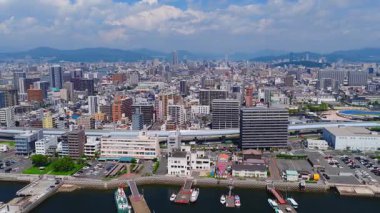 Hiroşima şehrinin havadan görünüşü şehir merkezindeki binaları gösteriyor. Yükseltilmiş bir hat üzerinde hareket eden bir tren, rıhtımlar ve bulutların altındaki uzak dağlar..