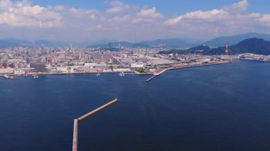 Hiroşima, Japonya 'nın havadan görünüşü rıhtımı, şehir ortası yükselen binaları, yeşil bir tepe ve bulutlu bir gökyüzünün altındaki uzak dağları gösteriyor..