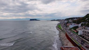 Japonya 'daki Kamakuro kasabasının kıyı yolu, okyanus dalgaları, Enoshima Adası' nda bir deniz feneri ve kıyı şeridi boyunca yavaş bir tepsisi olan hava manzarası..