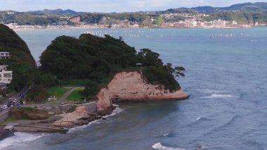 Japonya 'daki Kamakuro kasabasının hava manzarası yeşillik, dalgalar, sörfçüler, dağınık binalar, yüksek bir kule ve küçük botlarla kayalık bir uçurum gösteriyor..