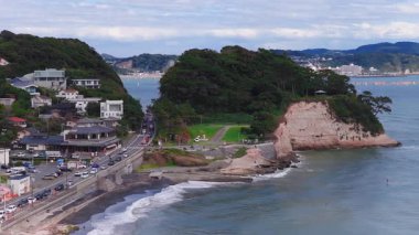 Japonya 'nın Kamakuro kentinin hava manzarası kayalık kayalıkları, tekneleri ve kürekleri olan sakin mavi denizi ve arka planda tepeleri olan uzak bir kasabayı gösteriyor..