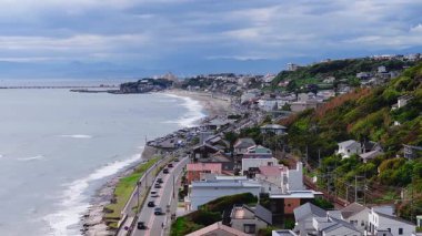 Japonya 'daki Kamakuro şehrinin hava görüntüsü Enoshima Adası ile bir köprü, hareket eden araçlar, sörfçüler, hafif dalgalar ve bulutlu bir gökyüzünün altındaki dağlar..