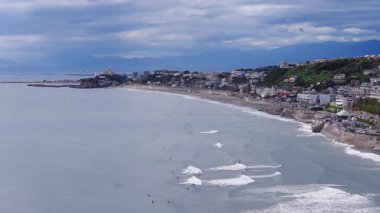 Japonya 'daki Kamakuro kasabasının hava görüntüsü, eğimli bir kıyı şeridi, okyanus dalgaları, sörfçüler, hareket eden araçlar ve bulutlu bir gökyüzünün altında bir köprü gösteriyor..