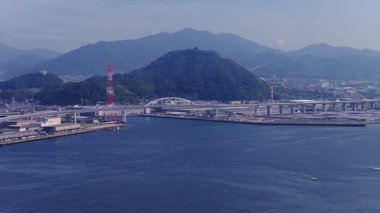 Japonya, Hiroşima 'nın hava görüntüsü, ahşap platformları olan bir nehir, kırmızı ve beyaz bir kule, bir köprü, ormanlık alanlar ve bir sanayi tesisi gösteriyor..